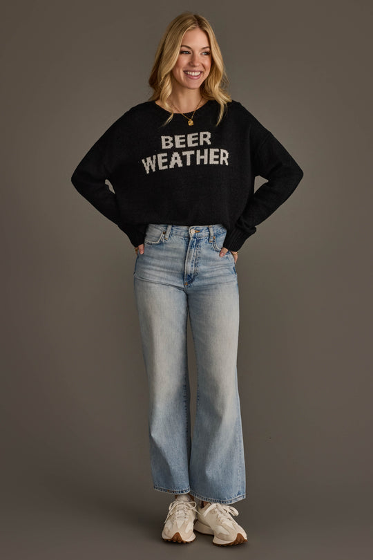 A smiling woman stands with her hands in her pockets, wearing Panache Apparel Collection's Beer Weather Lightweight Sweater in black, light blue high-waisted jeans, and white sneakers against a plain gray background.