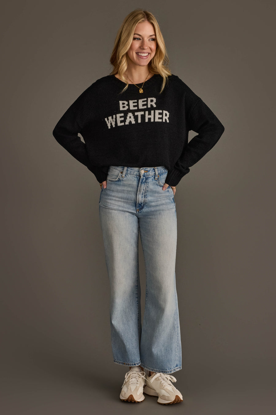 A smiling woman stands with her hands in her pockets, wearing Panache Apparel Collection's Beer Weather Lightweight Sweater in black, light blue high-waisted jeans, and white sneakers against a plain gray background.