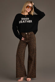 A woman smiles confidently, posing with one hand on her hip and the other adjusting her sunglasses, wearing Panache Apparel Collection’s Beer Weather Lightweight Sweater featuring the BEER WEATHER text, paired with brown leopard print wide-leg pants and heeled sandals.