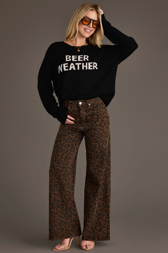 A woman smiles confidently, posing with one hand on her hip and the other adjusting her sunglasses, wearing Panache Apparel Collection’s Beer Weather Lightweight Sweater featuring the BEER WEATHER text, paired with brown leopard print wide-leg pants and heeled sandals.