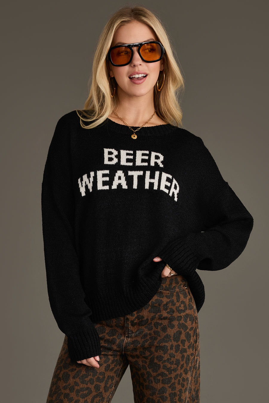 A woman stands against a neutral background, smiling with one hand on her hip. She wears large sunglasses, leopard-print pants, and the Panache Apparel Collection Beer Weather Lightweight Sweater, a cozy knit that adds a playful touch to her bold look.
