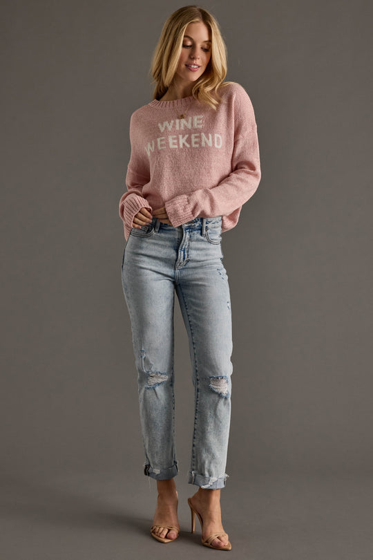 A woman smiles slightly, looking to the side, wearing the Panache Apparel Collection Light Pink Wine Weekend Lightweight Sweater with light blue ripped jeans and nude high-heeled sandals against a plain background.