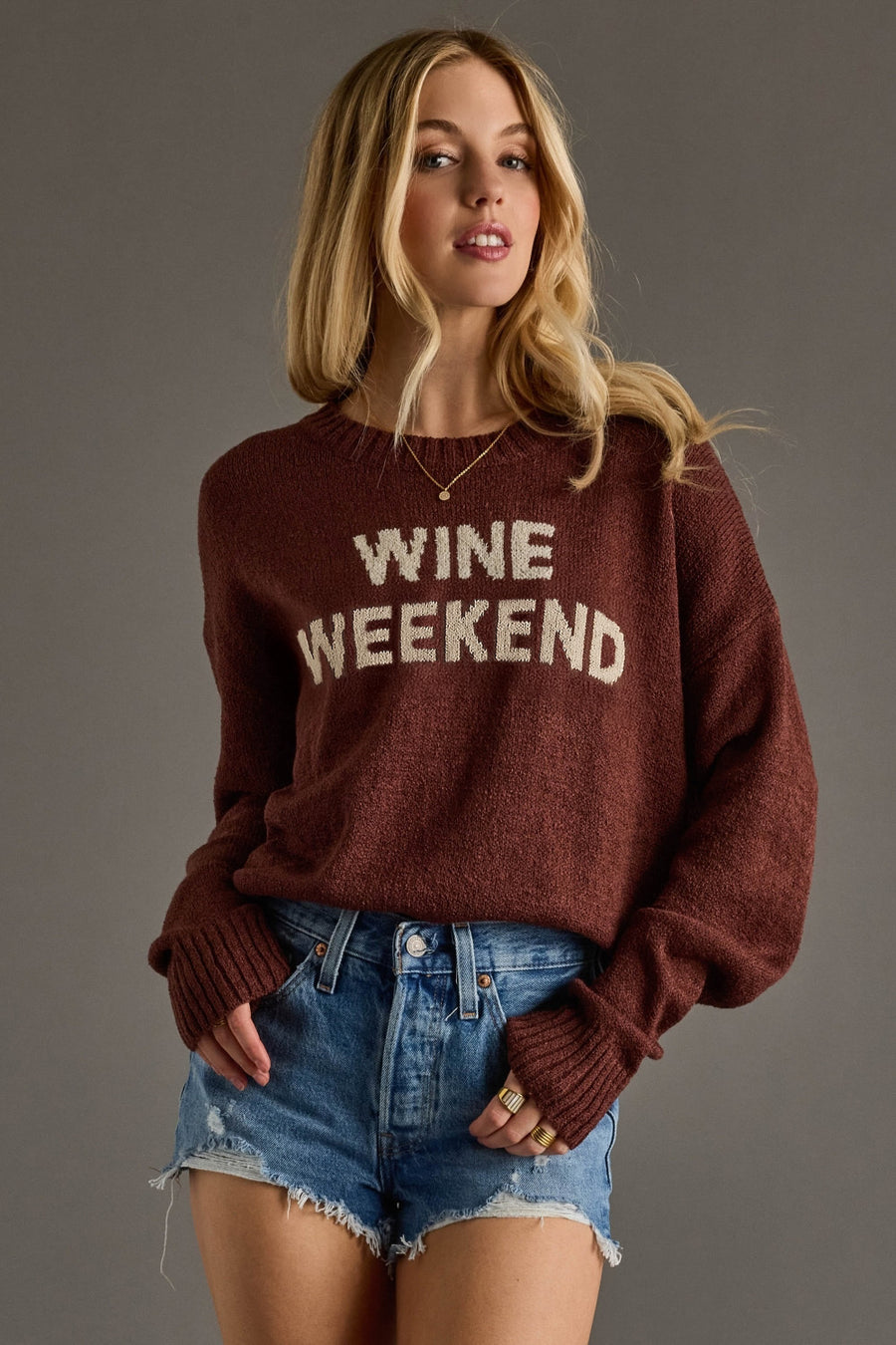 A woman with long blonde hair wears a Burgundy Wine Weekend Lightweight Sweater from Panache Apparel Collection and denim shorts, standing against a plain gray background, looking at the camera with a slight smile.