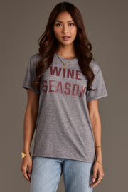 A person wearing a relaxed fit, gray Gray Wine Season Tee with maroon lettering, paired with blue jeans.