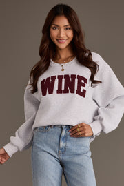 A smiling woman wears a Gray Wine Sweatshirt, an oversized light gray crewneck with burgundy chenille lettering, paired with light-wash blue jeans.