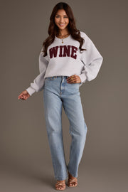 A woman wearing a Gray Wine Sweatshirt featuring oversized light gray fabric with WINE in maroon chenille lettering, paired with light-wash blue jeans.
