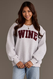 A woman wearing a Gray Wine Sweatshirt from Panache Apparel Collection, an oversized gray crewneck with the word WINE in burgundy, textured chenille lettering across the chest.