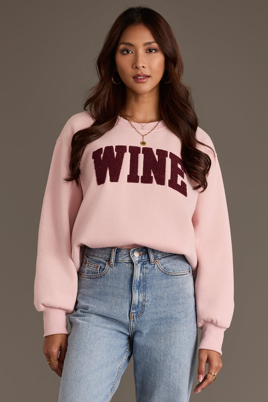 A person wearing a pale pink Panache Apparel Collection Pink Wine Sweatshirt featuring the word WINE in dark maroon chenille lettering across the chest.