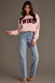 A smiling woman wears a pale pink oversized Pink Wine Sweatshirt from Panache Apparel Collection with WINE in dark chenille lettering across the chest, paired with light-wash blue jeans and strappy sandals.