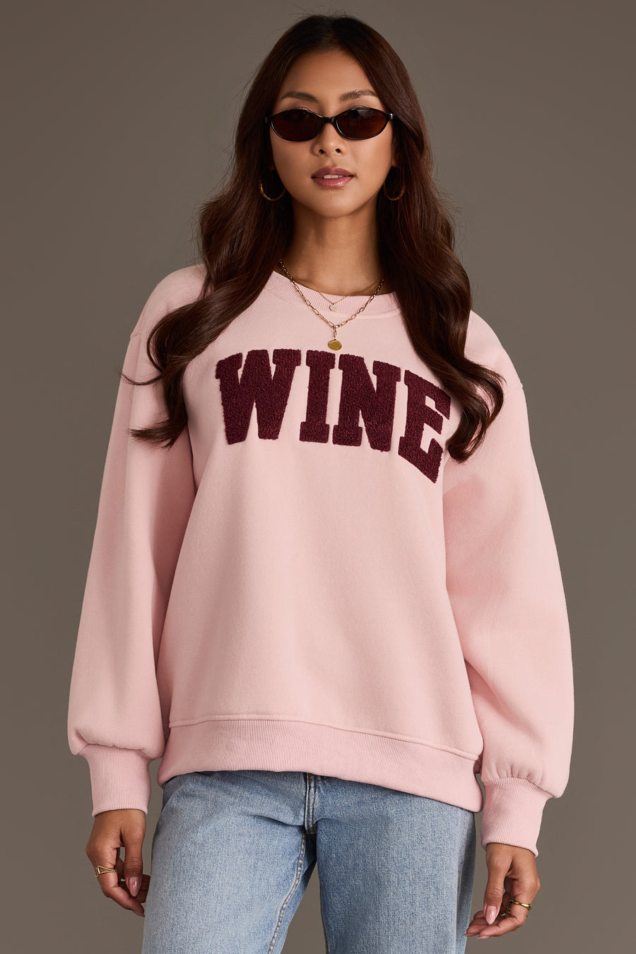 A person wearing a Panache Apparel Collection Pink Wine Sweatshirt, an oversized pink crewneck with the word WINE in dark red, textured chenille block letters across the chest.