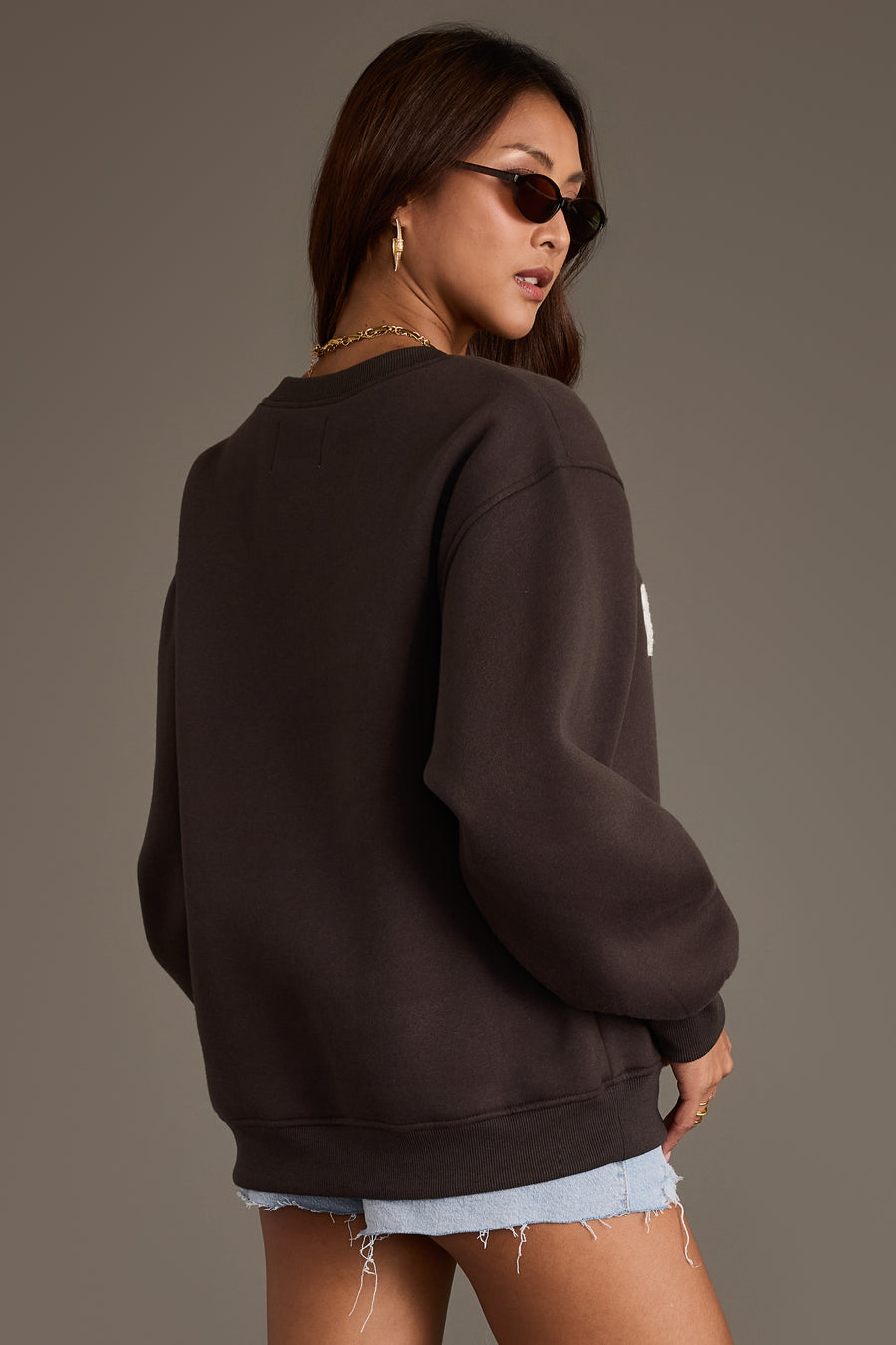 A woman stands sideways against a neutral background, looking over her shoulder while wearing the Panache Apparel Collection Beer Weather Sweatshirt in charcoal, paired with light denim shorts, sunglasses, and gold jewelry.