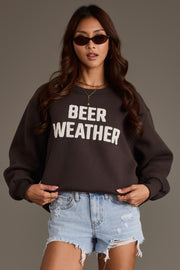 A woman wearing sunglasses and the black Beer Weather Sweatshirt from Panache Apparel Collection, crafted from soft fleece, stands against a plain background with her hands in her pockets. She pairs the cozy top with distressed denim shorts for a relaxed look.
