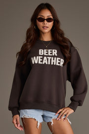 A woman wearing sunglasses and ripped denim shorts stands confidently against a plain background, dressed in the black, soft fleece Beer Weather Sweatshirt from Panache Apparel Collection.