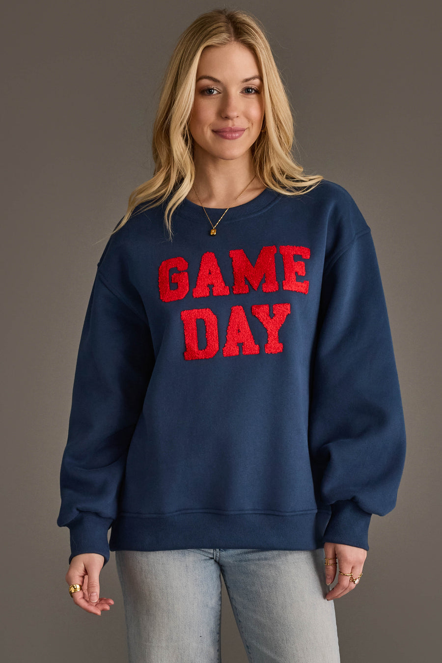 A woman models the Panache Apparel Collection Navy & Red Game Day Sweatshirt, featuring bold red "GAME DAY" lettering, as she smiles against a simple gray backdrop with her hands relaxed at her sides.