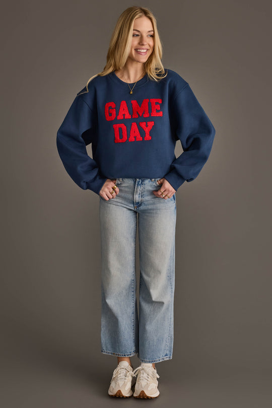 A smiling woman stands against a plain gray background, wearing the Panache Apparel Collection Navy & Red Game Day Sweatshirt, paired with light blue wide-leg jeans and white sneakers.