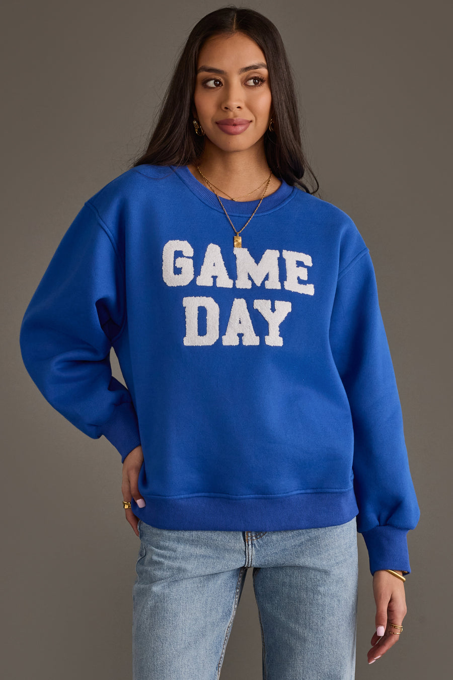 A woman wearing the Panache Apparel Collection's Royal Blue & White Game Day Sweatshirt, paired with light blue jeans and gold jewelry, stands against a plain gray background.