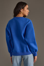 A woman stands facing away, wearing the Panache Apparel Collection Royal Blue & White Game Day Sweatshirt, light blue jeans, and gold hoop earrings against a neutral brown background.