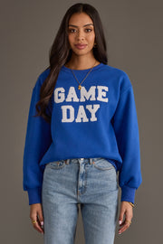 A woman with long dark hair wears the Panache Apparel Collection Royal Blue & White Game Day Sweatshirt paired with light-wash jeans, standing against a gray background.
