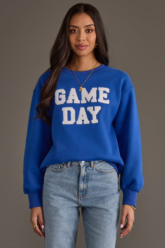 A woman with long dark hair wears the Panache Apparel Collection Royal Blue & White Game Day Sweatshirt paired with light-wash jeans, standing against a gray background.