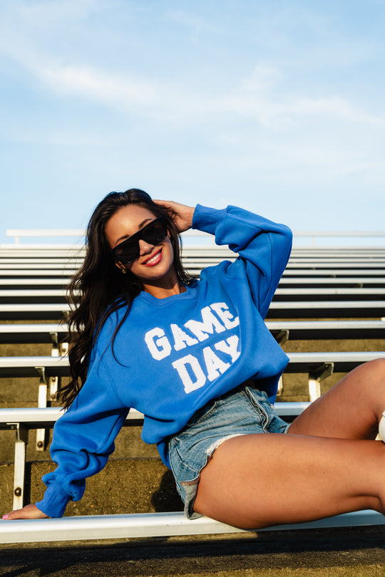 A woman wearing sunglasses and the Panache Apparel Collection Royal Blue & White Game Day Sweatshirt sits on the bleachers, smiling and leaning back with one hand in her hair under a sunny, clear sky.