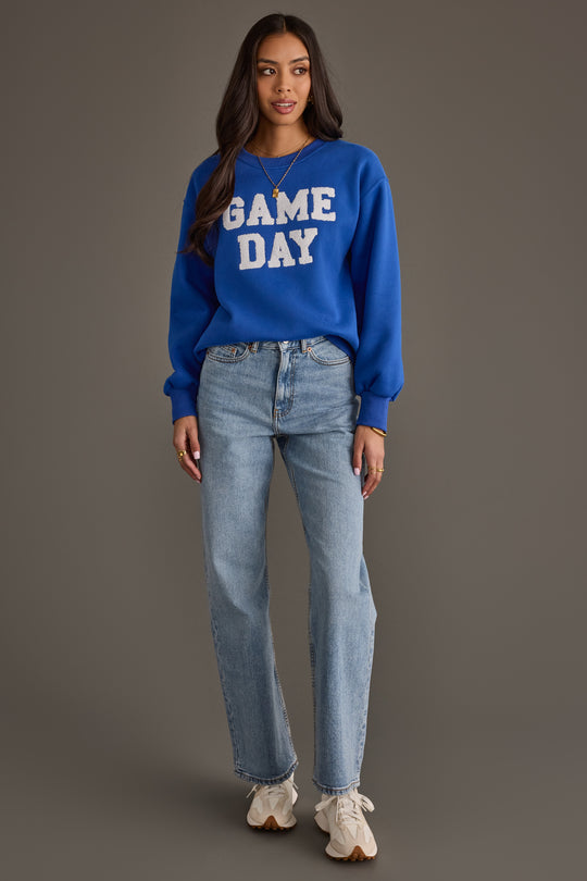 A woman wearing the Royal Blue & White Game Day Sweatshirt by Panache Apparel Collection, paired with light blue jeans and white sneakers, stands against a neutral background, looking at the camera with her arms relaxed at her sides.