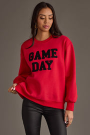 A woman models the Red & Black Game Day Sweatshirt from Panache Apparel Collection, featuring bold black "GAME DAY" lettering. She pairs it with black leather pants and gold hoop earrings, standing against a gray background and gazing slightly to the side.