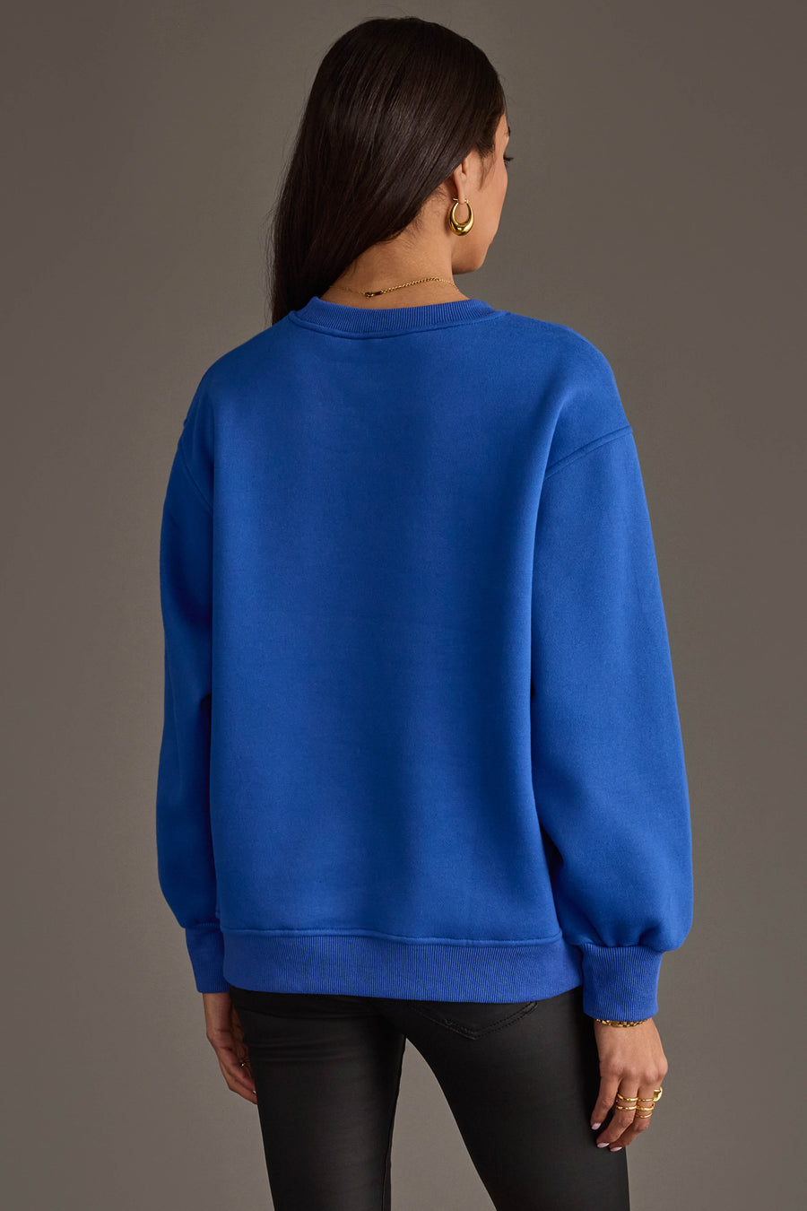 A woman with long brown hair, wearing gold hoop earrings and black pants, stands facing away from the camera against a plain gray background. She is wearing the Royal Blue & Red Game Day Sweatshirt by Panache Apparel Collection.