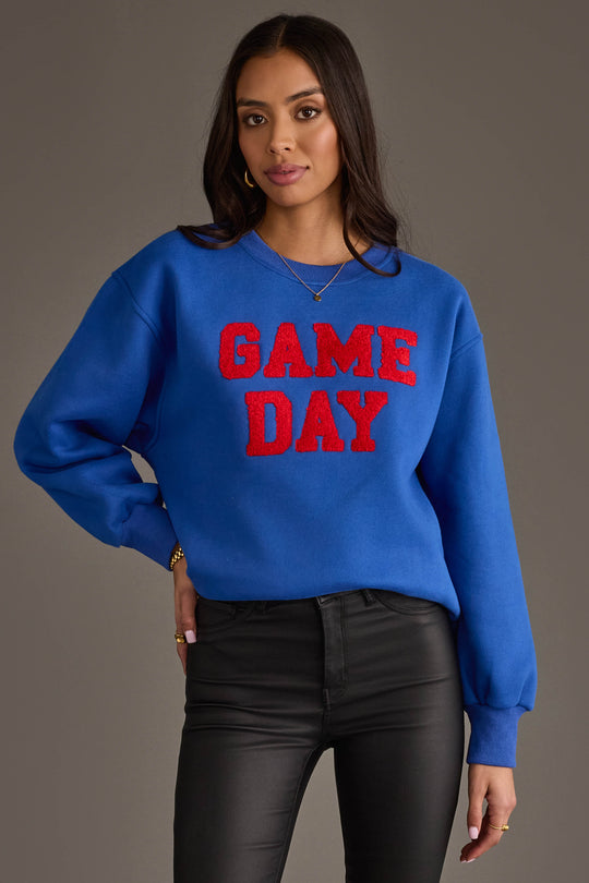 A woman models the Panache Apparel Collection’s Royal Blue & Red Game Day Sweatshirt, featuring bold red “GAME DAY” lettering, styled with black pants and gold jewelry against a neutral background.