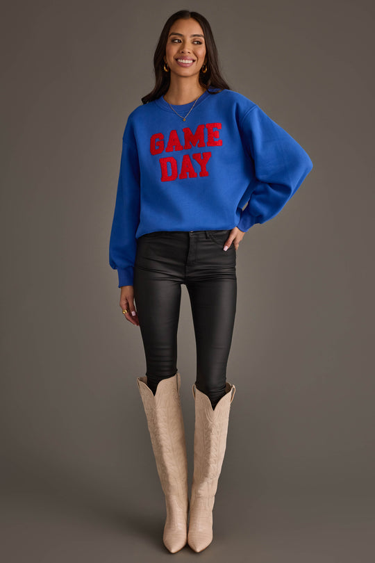 A smiling person is wearing a Panache Apparel Collection Royal Blue & Red Game Day Sweatshirt, paired with black skinny pants and cream cowboy boots, standing against a plain background.