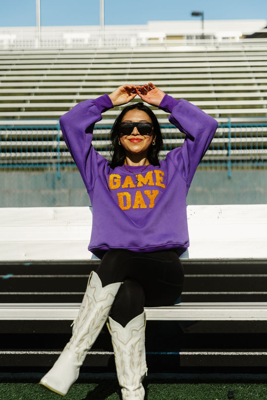 A woman sits on stadium bleachers, smiling with her arms raised above her head, wearing the Panache Apparel Collection Purple & Gold Game Day Sweatshirt, paired with black leggings, white cowboy boots, and sunglasses.