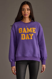 A woman stands facing forward against a gray background, wearing the Panache Apparel Collection Purple & Gold Game Day Sweatshirt featuring bold yellow "GAME DAY" lettering. She has long dark hair, gold earrings, and wears black pants.