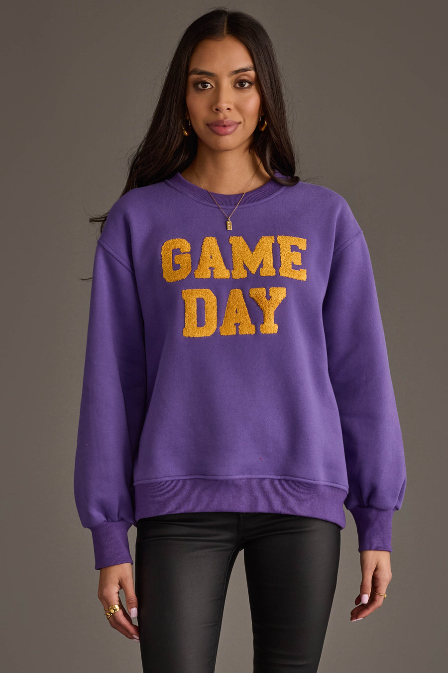 A woman stands facing forward against a gray background, wearing the Panache Apparel Collection Purple & Gold Game Day Sweatshirt featuring bold yellow "GAME DAY" lettering. She has long dark hair, gold earrings, and wears black pants.