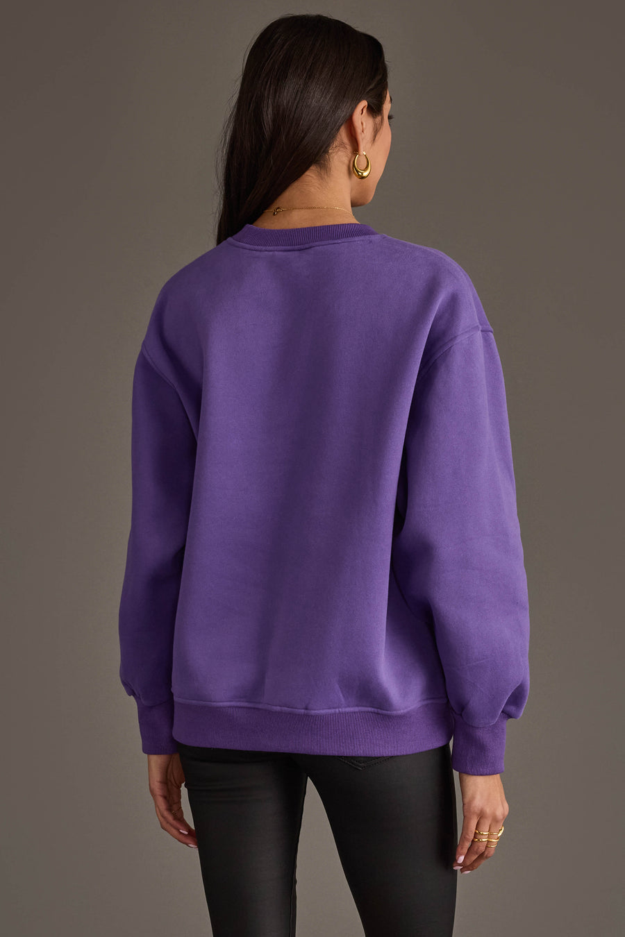 A woman with long dark hair, wearing gold hoop earrings and the Panache Apparel Collection’s Purple & Gold Game Day Sweatshirt, stands facing away from the camera against a plain gray background.
