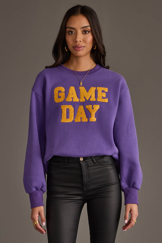 A woman wearing the Panache Apparel Collection's Purple & Gold Game Day Sweatshirt, featuring "GAME DAY" in bold yellow letters, paired with black pants and standing against a gray background.