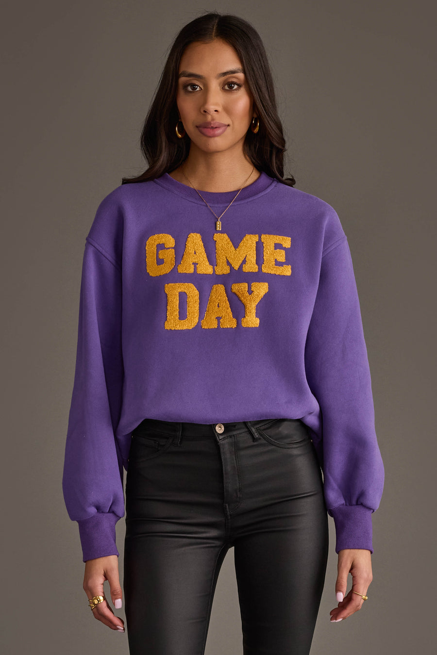 A woman wearing the Panache Apparel Collection's Purple & Gold Game Day Sweatshirt, featuring "GAME DAY" in bold yellow letters, paired with black pants and standing against a gray background.