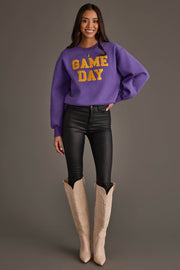 A woman stands and smiles against a plain gray background, wearing the Panache Apparel Collection’s Purple & Gold Game Day Sweatshirt with yellow lettering, paired with black pants and light-colored cowboy boots.