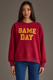 A model showcases the Panache Apparel Collection Maroon & Gold Game Day Sweatshirt, featuring textured gold "GAME DAY" lettering across the chest, paired with blue jeans against a neutral gray studio background.