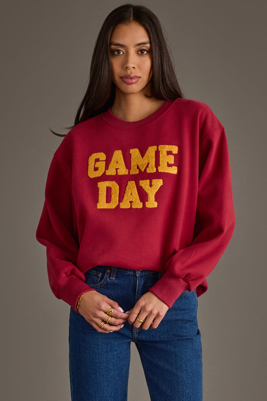 A woman stands against a plain background with her hands folded at her waist, wearing blue jeans and the Panache Apparel Collection’s Maroon & Gold Game Day Sweatshirt, featuring yellow "GAME DAY" lettering.