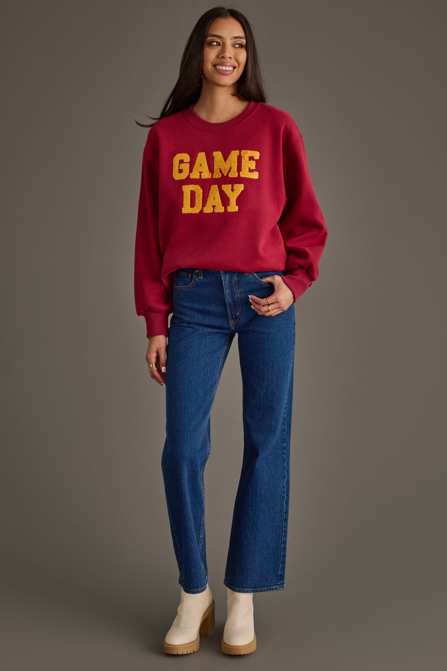 A woman stands smiling, wearing the Panache Apparel Collection’s Maroon & Gold Game Day Sweatshirt with yellow lettering, paired with blue jeans and beige boots against a plain gray background.