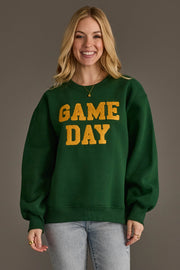A smiling woman with long blonde hair models the Panache Apparel Collection's Green & Gold Game Day Sweatshirt, featuring bold yellow "GAME DAY" lettering. She pairs it with light jeans and a gold pendant necklace while standing against a plain brown backdrop.