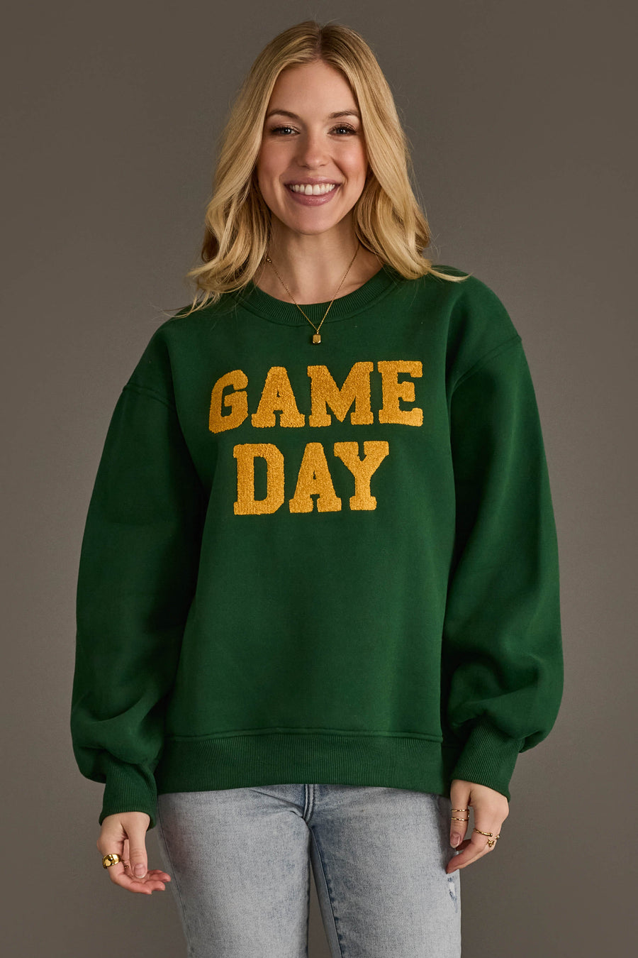 A smiling woman with long blonde hair models the Panache Apparel Collection's Green & Gold Game Day Sweatshirt, featuring bold yellow "GAME DAY" lettering. She pairs it with light jeans and a gold pendant necklace while standing against a plain brown backdrop.