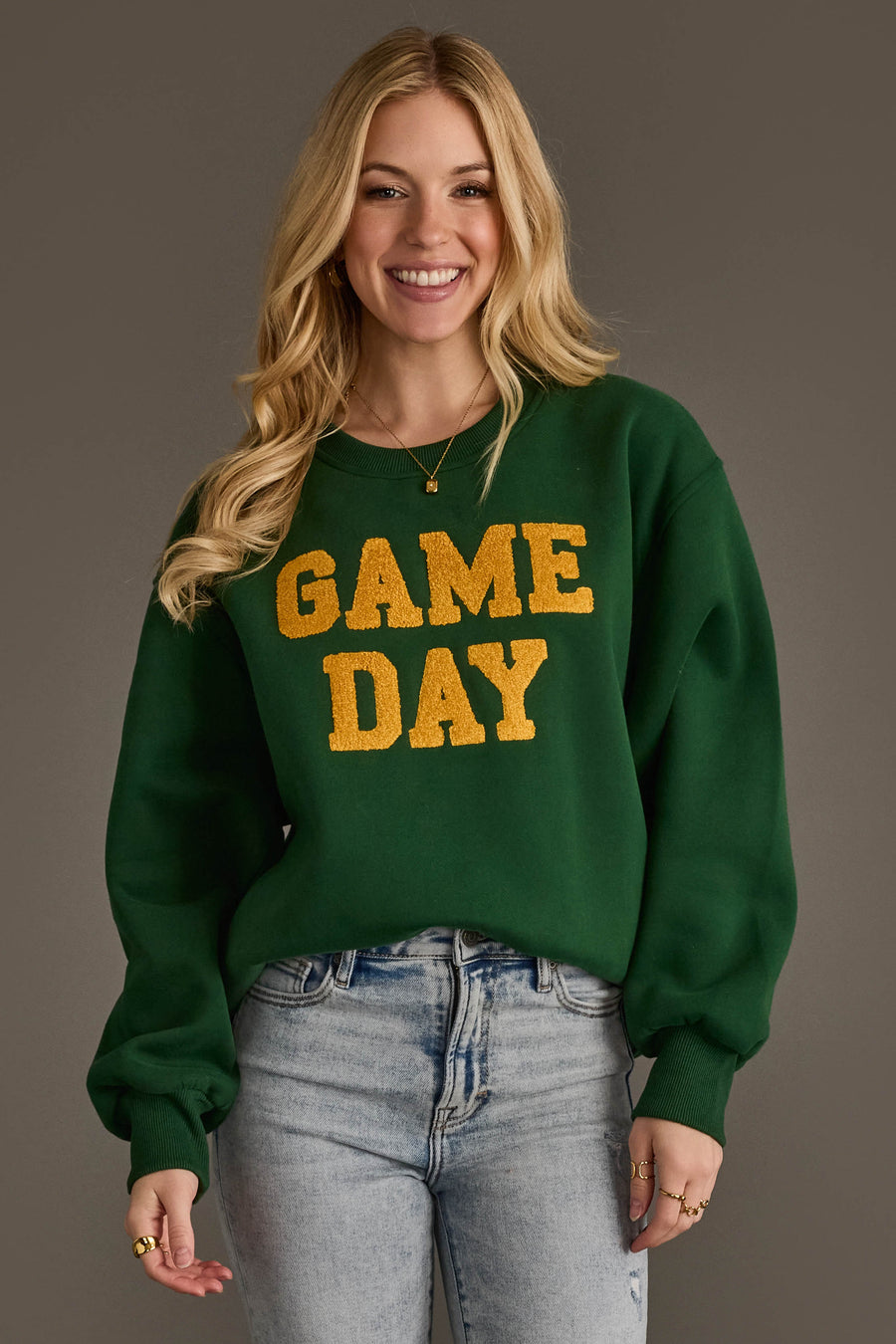 A smiling woman with long blonde hair models the Panache Apparel Collection's Green & Gold Game Day Sweatshirt, featuring yellow "GAME DAY" lettering, paired with light blue jeans and set against a plain gray background.
