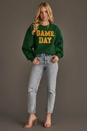 A woman in a Panache Apparel Collection Green & Gold Game Day Sweatshirt, ripped light blue jeans, and nude heels stands smiling against a plain brown background.