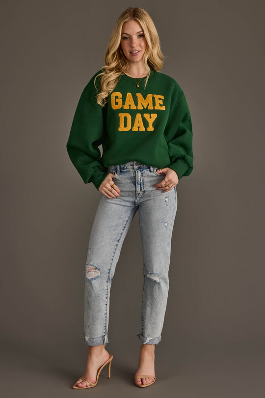 A woman in a Panache Apparel Collection Green & Gold Game Day Sweatshirt, ripped light blue jeans, and nude heels stands smiling against a plain brown background.
