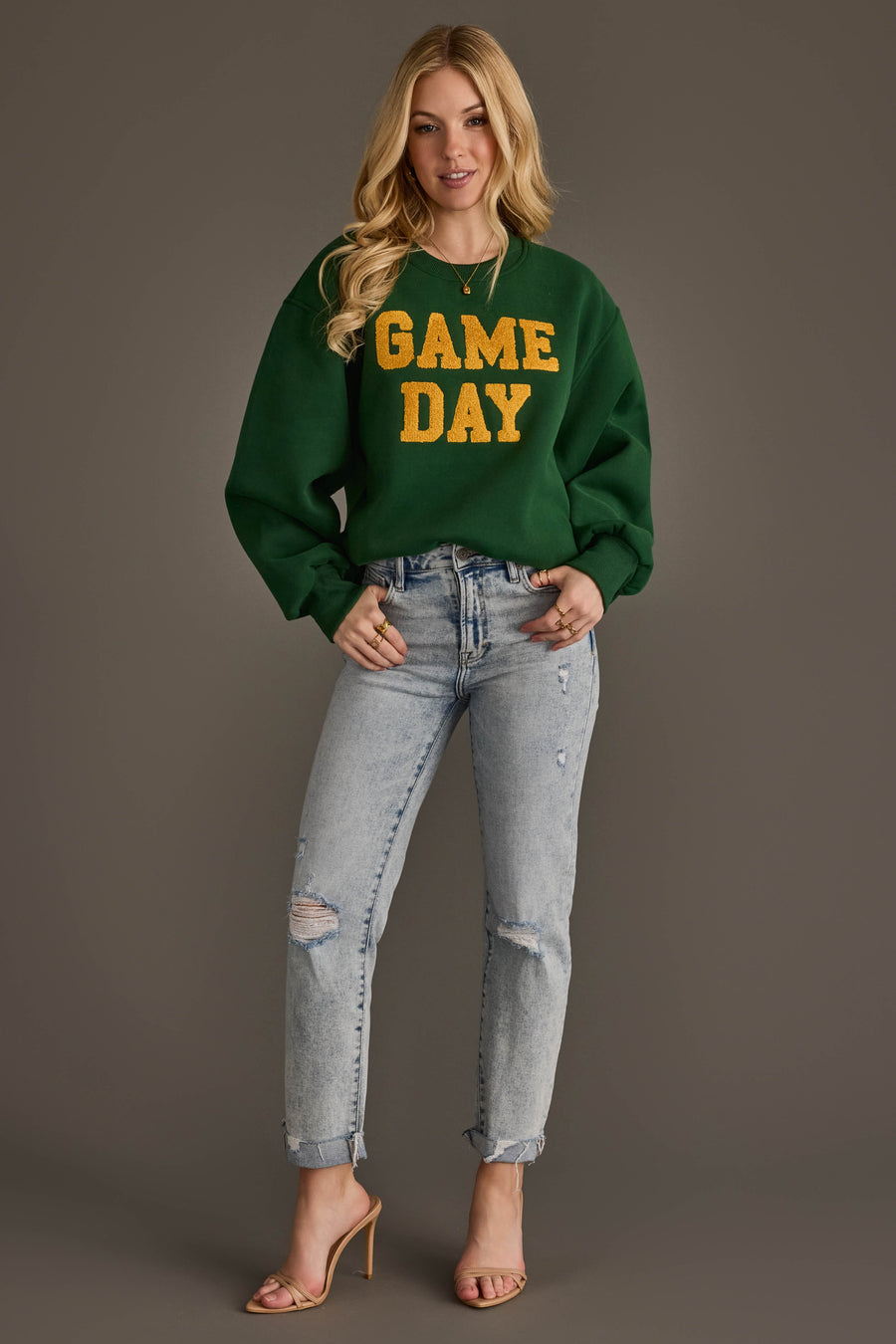 A woman in a Panache Apparel Collection Green & Gold Game Day Sweatshirt, ripped light blue jeans, and nude heels stands smiling against a plain brown background.