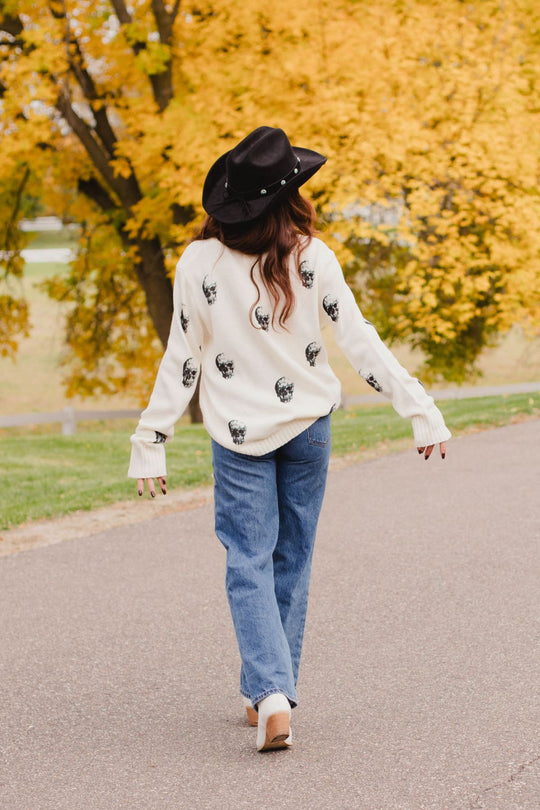 A person wearing a black cowboy hat and an Ivory Skull Sweater from the Panache Apparel Collection, paired with blue jeans, walking away on a paved path.