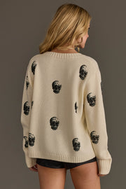 A woman with long blonde hair wears the Panache Apparel Collection Ivory Skull Sweater, featuring a black skull print, paired with black shorts as she stands against a plain background, facing away from the camera.