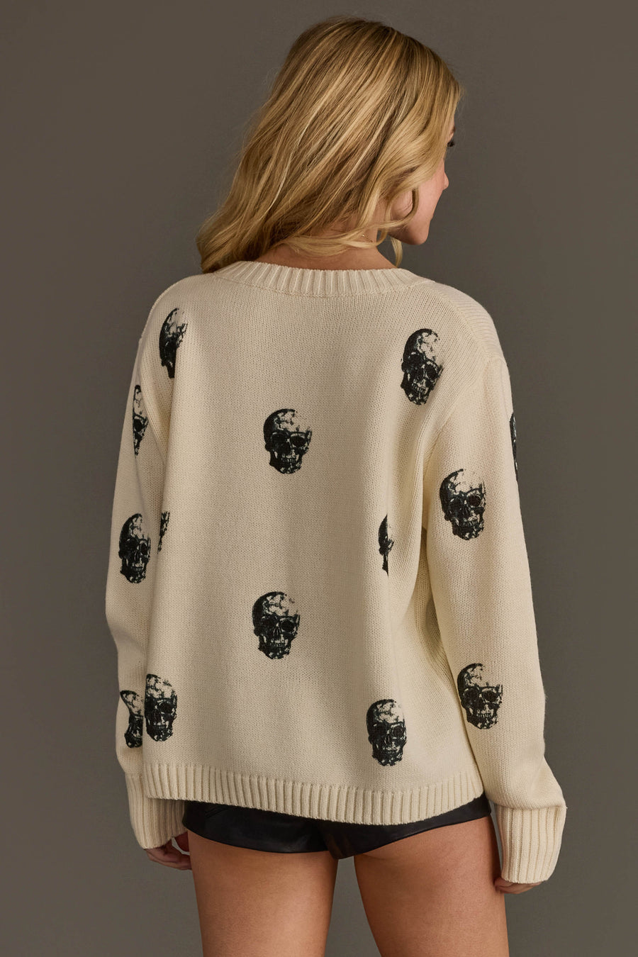 A woman with long blonde hair wears the Panache Apparel Collection Ivory Skull Sweater, featuring a black skull print, paired with black shorts as she stands against a plain background, facing away from the camera.