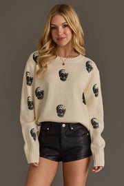 A woman with long blonde hair wears the Panache Apparel Collection’s Ivory Skull Sweater, a cream sweater featuring black skull prints, paired with black leather shorts, standing against a plain dark background.