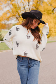 A person wearing a black cowboy hat and an Ivory Skull Sweater from Panache Apparel Collection featuring a black skull pattern.